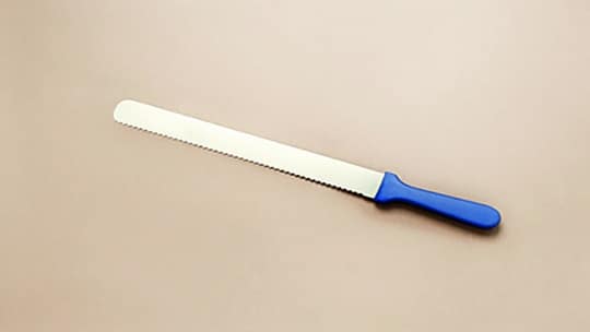 Ein langes gezacktes Messer mit blauem Griff liegt auf einer einfarbig beigen Fläche.