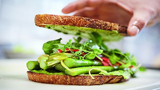 Eine Nahaufnahme einer Hand, die ein frisches Gemüsesandwich mit Blattgemüse, Spargel und buntem Gemüse zwischen Scheiben Vollkornbrot zusammenstellt.