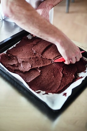 Eine Person verteilt den Schokoladenteig mit einem roten Spachtel gleichmäßig auf einem mit Pergament ausgelegten Backblech.
