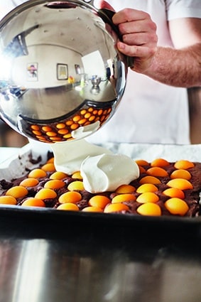 Eine Person gießt eine weiße, cremige Substanz aus einer Metallschüssel auf ein Backblech, das mit rohem Eigelb und einer dunklen Unterlage, möglicherweise Brownie-Teig, belegt ist. In der Schüssel spiegeln sich das Eigelb und die Küchenumgebung.