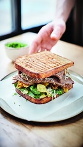 Eine Hand greift nach einem gegrillten Sandwich, das mit Aufschnitt, Salat, Sprossen und Kartoffelchips gefüllt ist und auf einem Teller mit einer kleinen Schale mit Grünzeug im Hintergrund serviert wird.