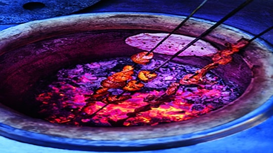 Metallspieße mit mariniertem Fleisch und Gemüse werden in einem traditionellen Tandoor-Ofen über glühenden Kohlen gebraten, die ein leuchtendes violettes und orangefarbenes Licht verbreiten.