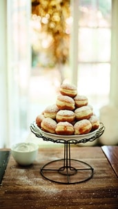 Eine Pyramide aus Puderzucker-Donuts ist auf einem dekorativen Kuchenständer gestapelt, daneben steht eine Schale mit Sahne auf einem Holztisch. Im Hintergrund sind ein sonnenbeschienenes Fenster und verschwommene goldene Lichter zu sehen.