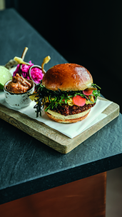 Ein Gourmet-Burger mit Blattgemüse und eingelegtem Gemüse wird auf einem Holzbrett mit Soße und bunt eingelegtem Gemüse auf einer dunklen Arbeitsplatte serviert.