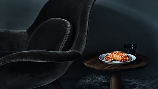Ein dunkler Plüschsessel neben einem kleinen hölzernen Beistelltisch mit einem Teller Croissants und einem Glas eines dunklen Getränks, in einem schwach beleuchteten Raum mit gemütlicher, stimmungsvoller Atmosphäre.
