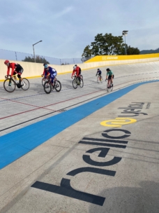 Radfahrer fahren auf einer steil abfallenden Radrennbahn im Freien, sie tragen Helme und Rennkleidung. Der Schriftzug "Hello Yellow" ist auf den Rand der Bahn in der Nähe der Kamera gemalt. Im Hintergrund sind Bäume und ein Zaun zu sehen.