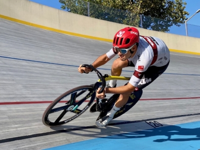 Ein Radfahrer, der einen roten Helm und ein weißes Trikot trägt, fährt mit hoher Geschwindigkeit auf einer abschüssigen Radrennbahn im Freien und neigt sich scharf in eine Kurve.