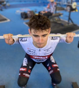 Ein junger Mann in Sportkleidung hockt mit einer Langhantel auf den Schultern in einem Fitnessstudio mit blauem Bodenbelag und konzentriert sich während seines Trainings. Andere Fitnessgeräte und Menschen erscheinen unscharf im Hintergrund.