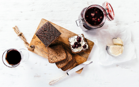 Scheiben von dunklem Brot auf einem Holzbrett, eine davon mit Weichkäse und Beerenkonfitüre belegt. Daneben stehen ein Glas mit Beerenmarmelade, eine Scheibe Weichkäse auf Pergament und ein Glas Rotwein auf einer weißen Unterlage.