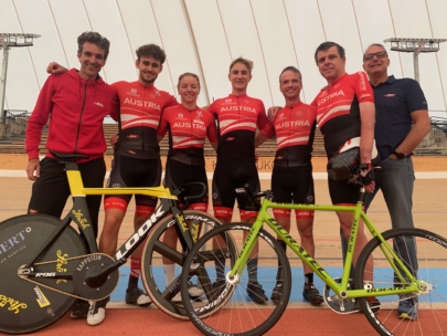 Sechs Personen stehen auf einer Indoor-Radrennbahn, fünf in passenden roten Austria-Radtrikots und eine in einem blauen Hemd. Zwei Rennräder stehen vor der Gruppe. Alle lächeln.