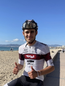Ein Radfahrer in weißem und kastanienbraunem Trikot und Helm zeigt lächelnd zwei Daumen nach oben, während er an einem Sandstrand mit blauem Himmel und fernen Gebäuden im Hintergrund steht.