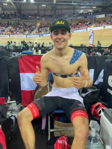 Ein lächelnder männlicher Radfahrer in schwarz-roten Austria-Shorts, der ohne Hemd und mit blauem Klebeband auf der Schulter sitzt, zeigt zwei Daumen nach oben bei einer Indoor-Velodrom-Veranstaltung, mit Zuschauern und Fahrradmarken im Hintergrund.