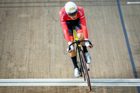 Ein Radfahrer in einem rot-schwarzen Rennanzug und mit weißem Helm fährt auf einem Bahnrad in einem Indoor-Velodrom, aufgenommen von oben.
