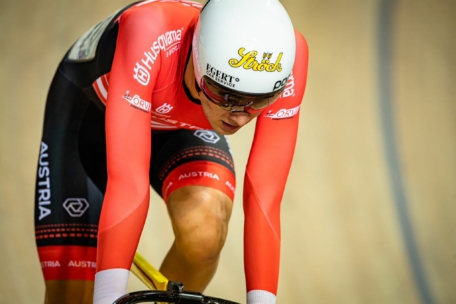 Ein Radfahrer in einem roten Austria-Trikot und einem weißen Helm fährt auf einer Indoor-Bahn. Der Kopf des Sportlers ist nach unten gerichtet, und auf Helm und Trikot sind Sponsorenlogos zu sehen.