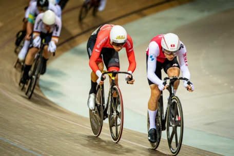 Zwei Radsportler in aerodynamischen Helmen und Rennanzügen biegen auf einer Indoor-Velodrombahn in eine Kurve ein und fahren dicht hintereinander, gefolgt von zwei weiteren Radfahrern.