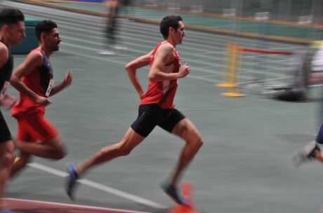Drei männliche Athleten in roten Uniformen laufen auf einer Indoor-Bahn, wobei einer die Gruppe anführt. Der Hintergrund erscheint unscharf, was ihre Geschwindigkeit und Bewegung unterstreicht.
