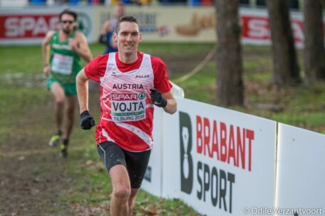 Ein männlicher Läufer in einem rot-weißen Österreich-Trikot nimmt lächelnd an einem Crosslauf teil, ein anderer Läufer folgt ihm. Die Strecke ist grasbewachsen mit Bäumen und einem Brabant Sport-Schild auf der rechten Seite.