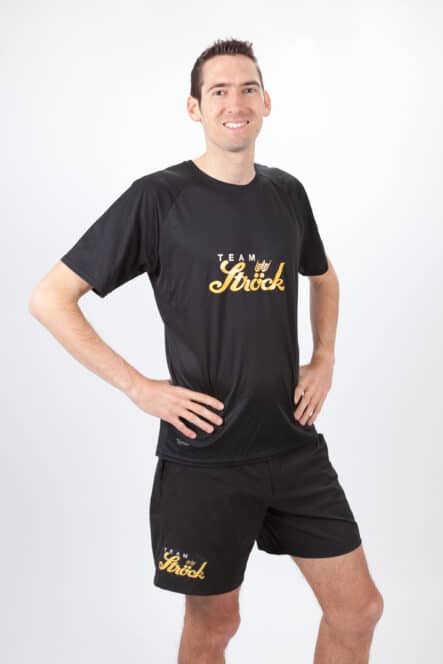 Ein lächelnder Mann steht mit den Händen in den Hüften, trägt ein schwarzes Sport-T-Shirt und Shorts mit der gelben Aufschrift Team My Stroke auf beiden Kleidungsstücken vor einem schlichten weißen Hintergrund.