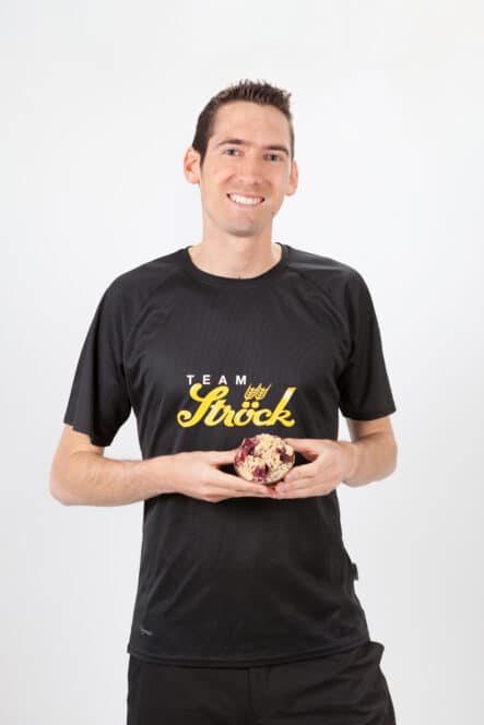 Ein lächelnder Mann in einem schwarzen Team Ströck-T-Shirt hält einen Muffin mit Beeren in beiden Händen und steht vor einem schlichten weißen Hintergrund.