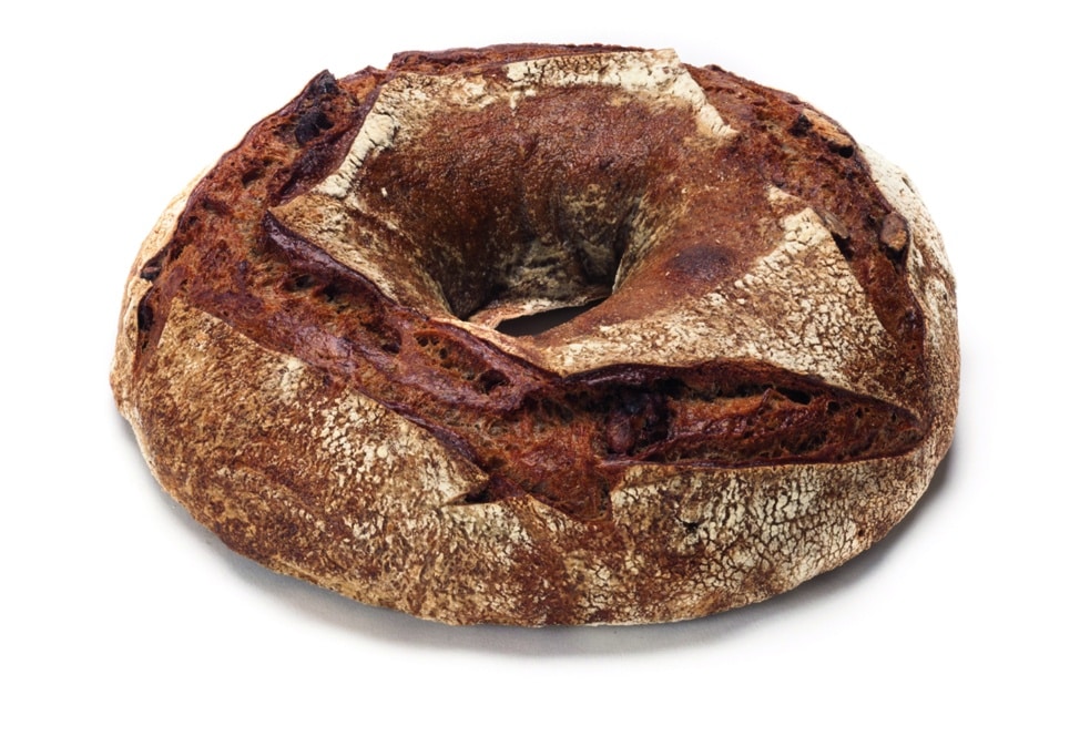 Ein runder, rustikaler Brotlaib mit einem Loch in der Mitte, mit einer dunklen, knusprigen Außenseite und tiefen Rillen auf der Oberfläche, vor einem weißen Hintergrund.