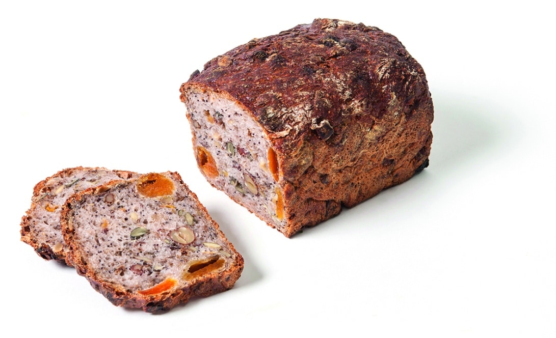 Ein Laib Mehrkornbrot mit Nüssen und Trockenfrüchten, ein Ende aufgeschnitten, um das dichte, strukturierte Innere zu enthüllen, und zwei Scheiben daneben, alles auf weißem Hintergrund.