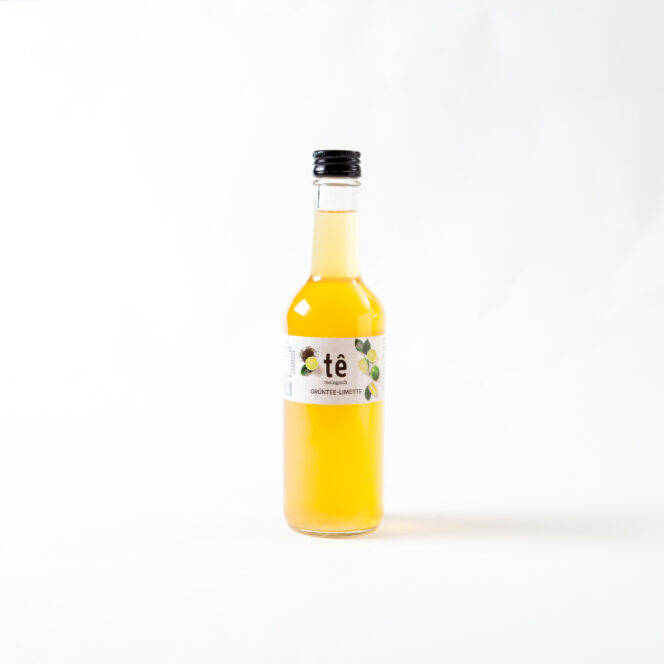 Eine mit gelber Flüssigkeit gefüllte Klarglasflasche mit schwarzem Schraubverschluss und weißem Etikett mit dem Wort "tè" und Abbildungen von Zitronen vor einem schlichten weißen Hintergrund.