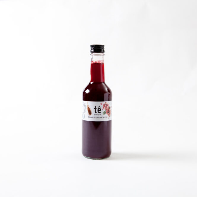 Eine mit dunkelroter Flüssigkeit gefüllte Klarglasflasche mit der Aufschrift "tê" und Abbildungen von Hibiskusblüten und Granatäpfeln steht vor einem schlichten weißen Hintergrund.