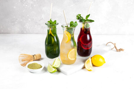 Drei Gläser mit Strohhalmen, gefüllt mit grünen, gelben und roten Getränken, garniert mit Zitronenscheiben und Minze, stehen auf einer weißen Tafel. In der Nähe befinden sich Limettenspalten, eine Zitrone, eine Schale mit grünem Pulver und ein Bambusbesen.