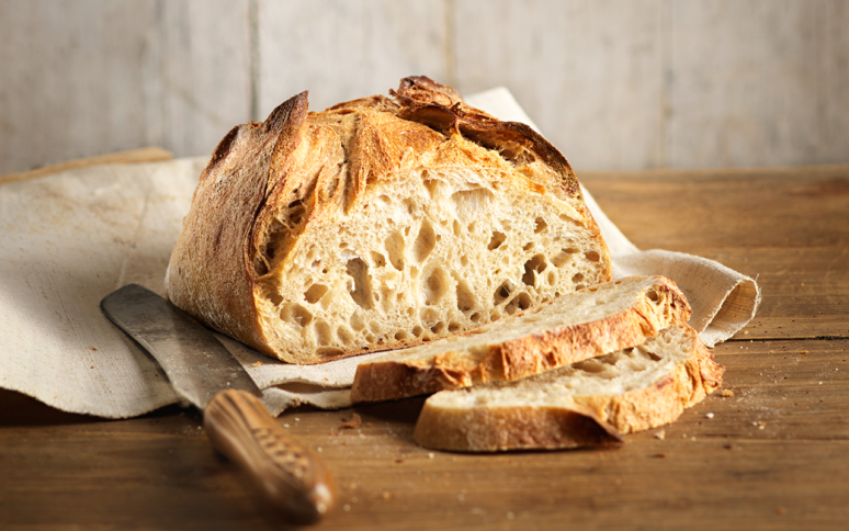 Auf einem Holztisch liegt auf einem Tuch ein rustikaler Laib handwerklich hergestellten Brotes, vor dem zwei Scheiben aufgeschnitten liegen. Ein Messer mit einem Holzgriff liegt daneben. Das Brot hat eine goldene, knusprige Oberfläche und eine luftige, offene Krume.