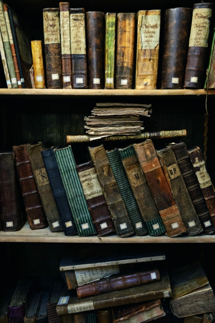 Ein Bücherregal, gefüllt mit alten, abgenutzten Büchern und Dokumenten, einige gestapelt, andere schräg angelehnt. Die Bücher haben verwitterte Einbände in verschiedenen Braun-, Grün- und Hellbrauntönen, die ein Gefühl von Alter und Geschichte vermitteln.