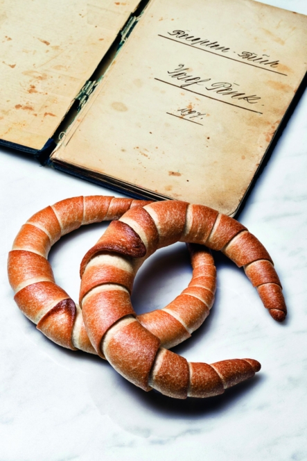 Zwei halbmondförmige Brötchen liegen auf einer weißen Fläche vor einem alten, aufgeschlagenen Rezeptbuch mit handschriftlichem Text auf vergilbten Seiten.