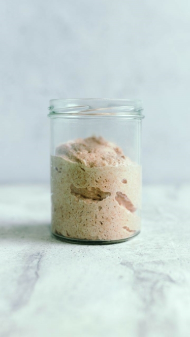 Ein Glasgefäß, das zur Hälfte mit blubberndem, hellbraunem Sauerteigstarter gefüllt ist, steht auf einer weißen, marmorierten Fläche vor einem hellen Hintergrund.