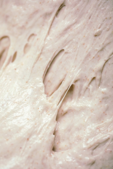 Nahaufnahme eines klebrigen, dehnbaren Brotteigs mit glatter, leicht glänzender Oberfläche und sichtbaren Luftblasen und Strängen. Der Teig hat eine hellbeige Farbe mit kleinen Flecken im Teig.