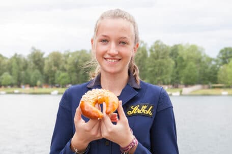 Eine junge Frau steht im Freien an einem See, lächelt und hält eine weiche, mit grobem Salz bestreute Brezel in der Hand. Sie trägt eine marineblaue Jacke mit aufgesticktem Schock. Im Hintergrund sind Bäume und Wasser zu sehen.
