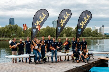 Eine Rudermannschaft posiert auf einem Steg an einem Fluss mit ihrem Boot und ihren Paddeln. Im Hintergrund sind drei große Schockflaggen zu sehen. Die Teammitglieder tragen passende marineblaue Hemden und lächeln. Stadtgebäude und Bäume sind zu sehen.