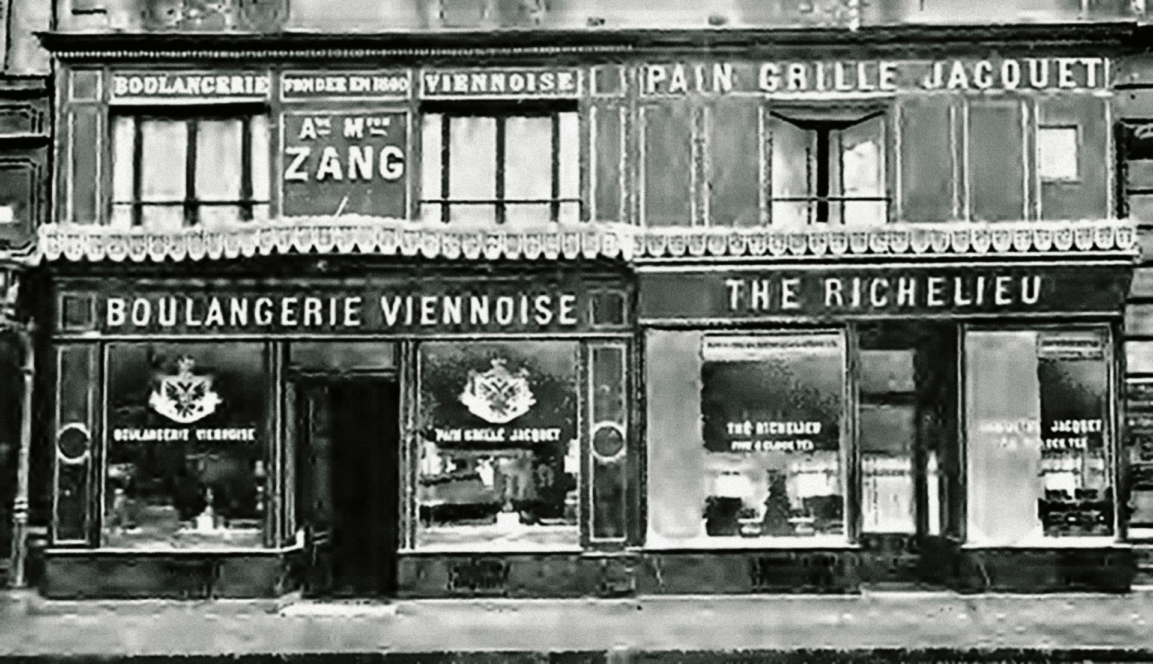 Ein altes Schwarz-Weiß-Foto einer Bäckerei in Frankreich mit Schildern wie Boulangerie Viennoise, Pain Grillé Jacquet und The Richelieu. Große Schaufenster zeigen Backwaren.