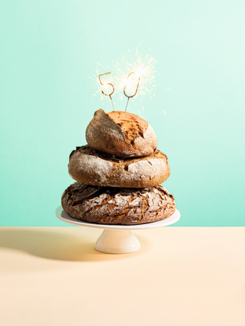 Ein Stapel von drei rustikalen Broten auf einem Kuchenständer mit Wunderkerzen in Form der Zahl 50 auf der Spitze vor einem mintgrünen Hintergrund.