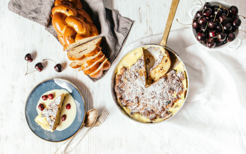 Eine runde Auflaufform mit einer mit Puderzucker bestreuten Scheibe Brotpudding, eine Portion auf einem Teller mit Kirschen und Vanillepudding, in Scheiben geschnittenes Zopfbrot, lose Kirschen und eine Schale mit Kirschen auf einem weißen Tisch.