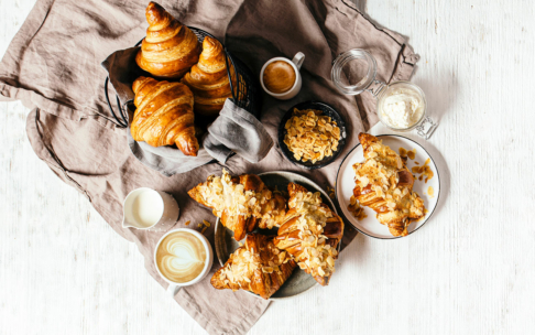 Ein Frühstücksbuffet mit Croissants und Mandelhörnchen auf Tellern und in einem Korb, Tassen mit Kaffee, Milch, einem Glas Sahne, Mandelblättchen und einem hellbraunen Tuch auf einem weißen Holztisch.