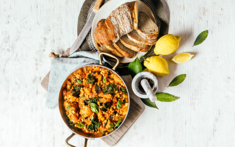 Auf einem weißen Holztisch steht ein Topf mit Risotto auf Tomatenbasis mit Grünzeug und Kräutern neben einem Teller mit rustikalem Brot in Scheiben, zwei ganzen Zitronen, einem Mörser und Stößel mit Pfefferkörnern und Lorbeerblättern.