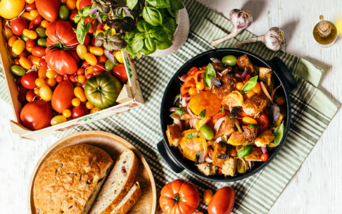 Ein Tisch mit einem Korb voller bunter Tomaten, einem runden Brotlaib mit Scheiben, einer Pfanne mit Panzanella-Salat mit Tomaten, Basilikum und Brotwürfeln, Knoblauchzwiebeln und einer Pflanze, von oben aufgenommen.