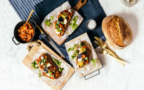 Drei belegte Brötchen mit Gemüse und Grünzeug auf Holztabletts, ein Topf mit Eintopf und ein Holzlöffel, ein runder Laib Brot, goldenes Geschirr und eine Schale mit Salz auf einer weiß-marine gestreiften Tischdecke.