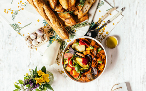 Eine Schüssel mit einem bunten Gemüseeintopf mit Kräutern steht auf einem weißen Tisch, umgeben von Baguettescheiben, einem Knoblauchbündel, einer kleinen Tasse Olivenöl und einem Strauß gelber Blumen.