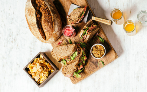 Ein Holzbrett mit aufgeschnittenen, mit Gemüse und Fleisch gefüllten Sandwiches, ein Laib rustikales Brot, eine Schale Senf, eingelegte Zwiebeln, eine Schale mit Pommes frites und zwei Gläser Orangensaft auf einer hellen Unterlage.