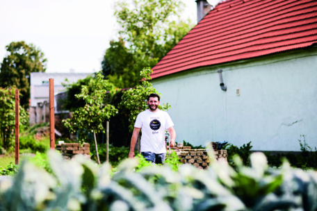 Ein Mann in einem weißen T-Shirt steht lächelnd in einem üppigen Garten mit grünen Pflanzen. Hinter ihm steht ein weißes Gebäude mit einem roten Dach und einigen Bäumen. Die Szene sieht hell und sonnig aus.