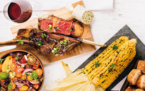 Ein gedeckter Tisch mit gegrilltem Steak auf einem Schneidebrett, Maiskolben mit Kräutern, einem bunten Tomatensalat, geschnittenem Brot, einer Runde Kräuterbutter und einem Glas Rotwein.