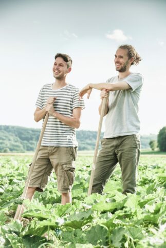 Zwei Männer stehen an einem sonnigen Tag auf einer grünen Wiese, lächeln und stützen sich auf Holzwerkzeuge. Beide tragen Freizeitkleidung, der eine ein gestreiftes Hemd, der andere ein schlichtes Hemd. Im Hintergrund sind üppige Pflanzen und Hügel zu sehen.