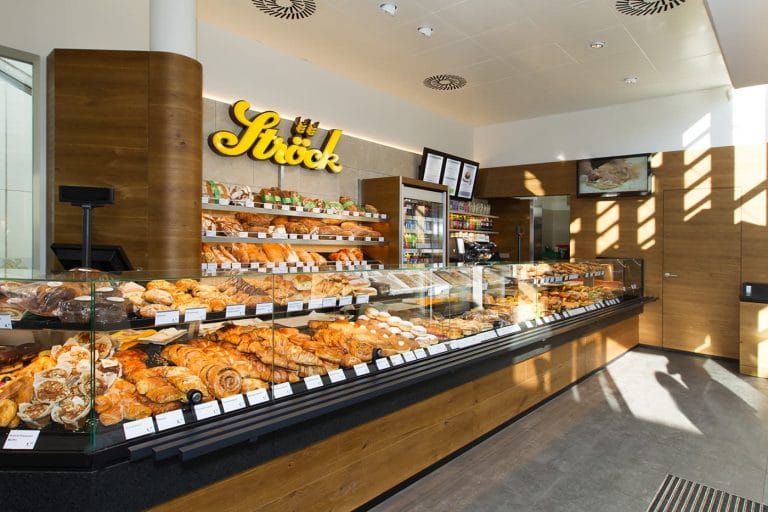 Eine moderne Bäckerei mit einer Glasvitrine, gefüllt mit verschiedenen Backwaren und Brot. Hinter der Theke befinden sich Holzregale mit weiteren Backwaren. Der Innenraum ist mit natürlichem Licht gut beleuchtet und trägt ein Schild mit der Aufschrift Ströck.