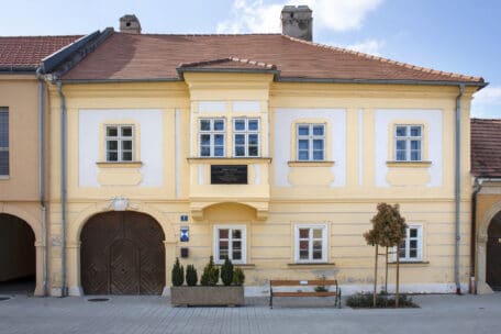 Ein historisches, gelbes, zweistöckiges Gebäude mit weißer Verkleidung und rotem Ziegeldach. Es hat mehrere Fenster, eine große hölzerne Bogentür, eine Tafel über dem Eingang, Topfpflanzen, eine Bank und einen kleinen Baum davor.