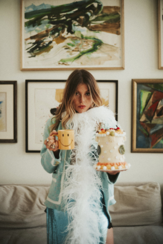 Eine Frau in hellblauem Outfit und weißer Federboa hält eine Smiley-Tasse und eine dekorierte Torte und steht vor einer Wand mit abstrakter Kunst.